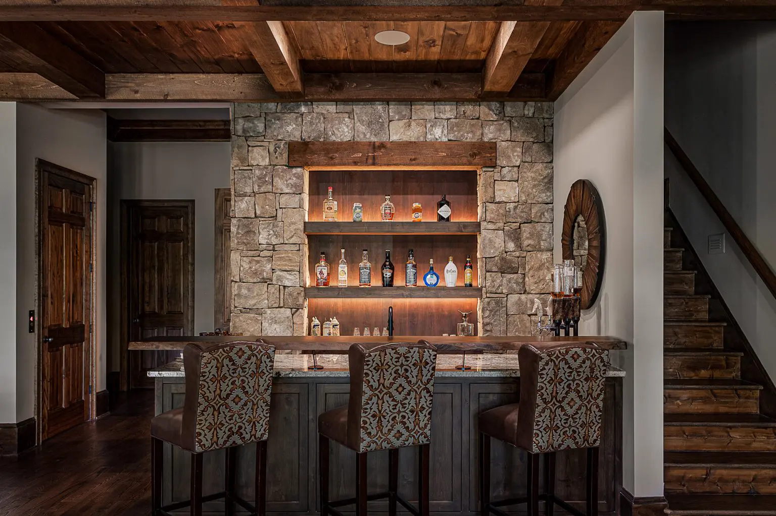 Home bar with stone accent wall, wooden shelves for liquor display, and patterned bar stools. Home bar with stone accent wall, wooden shelves for liquor display, and patterned bar stools.