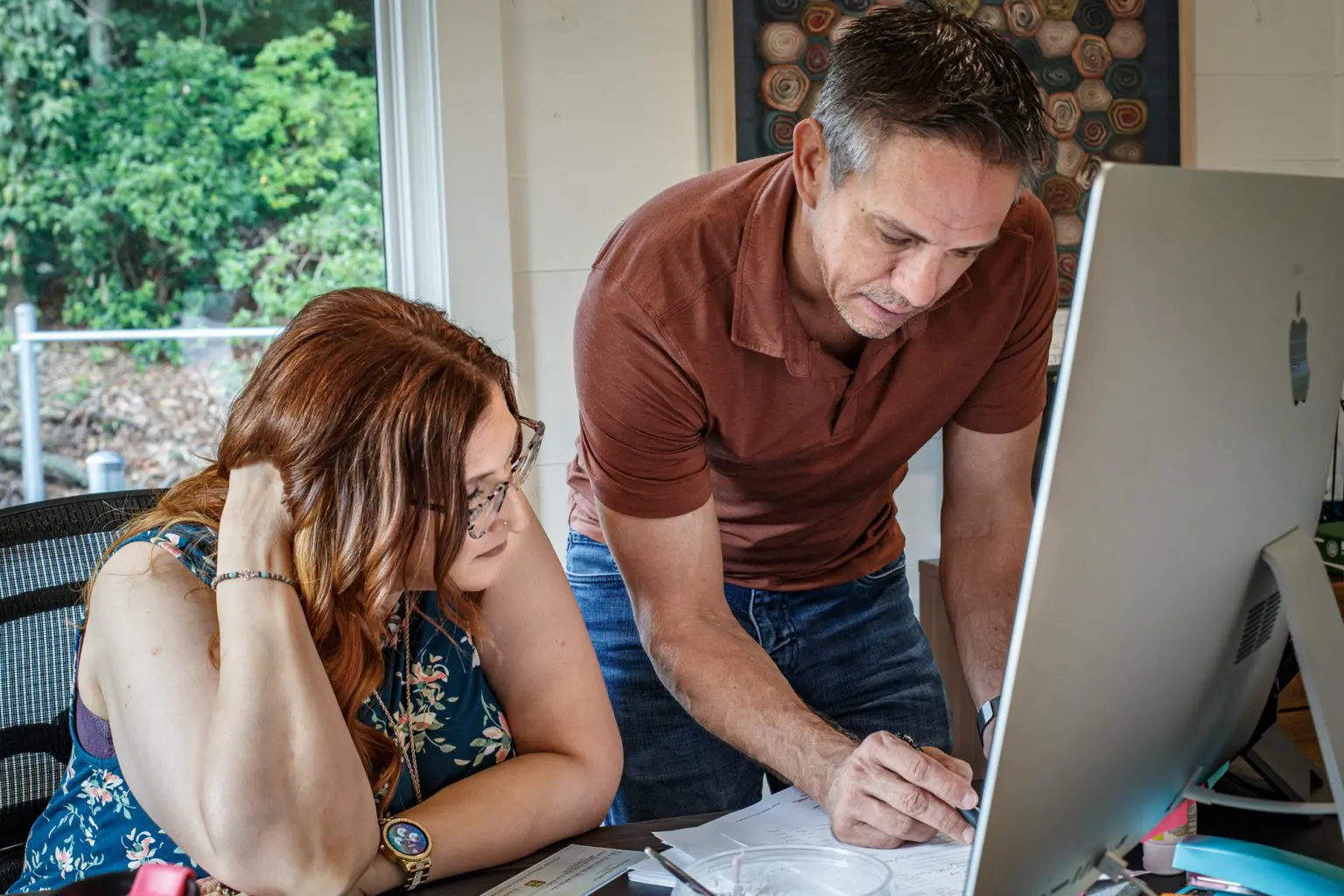 Two professionals collaborating at a computer desk with documents, in a well-lit room with greenery outside the window. Two professionals collaborating at a computer desk with documents, in a well-lit room with greenery outside the window.